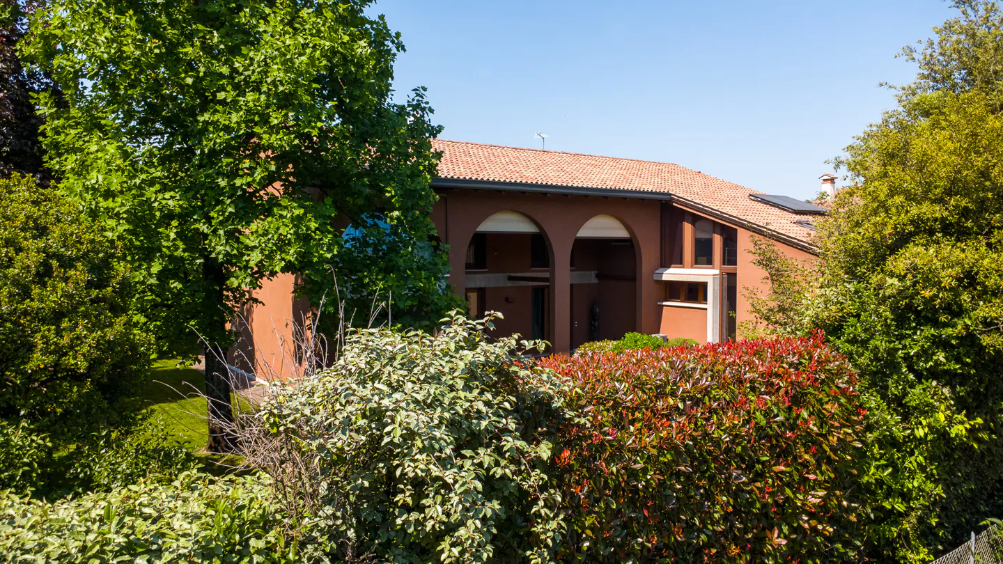 Exterior view of a terracotta-colored house with arched openings and a red tile roof, surrounded by lush green trees and bushes.