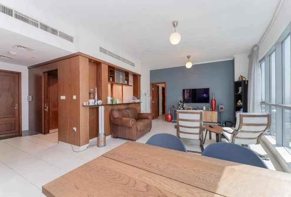 Modern living room with a wooden dining table, brown leather sofa, two white armchairs, and a blue accent wall with a TV.