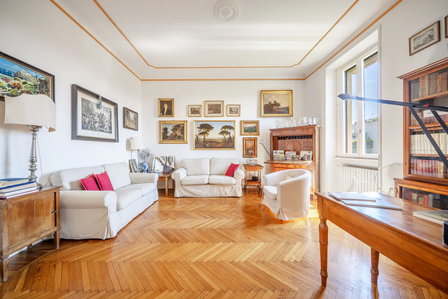 Bright living room with white sofas, wood floors, and many framed paintings on the wall. A wooden desk and bookcase are near the window.