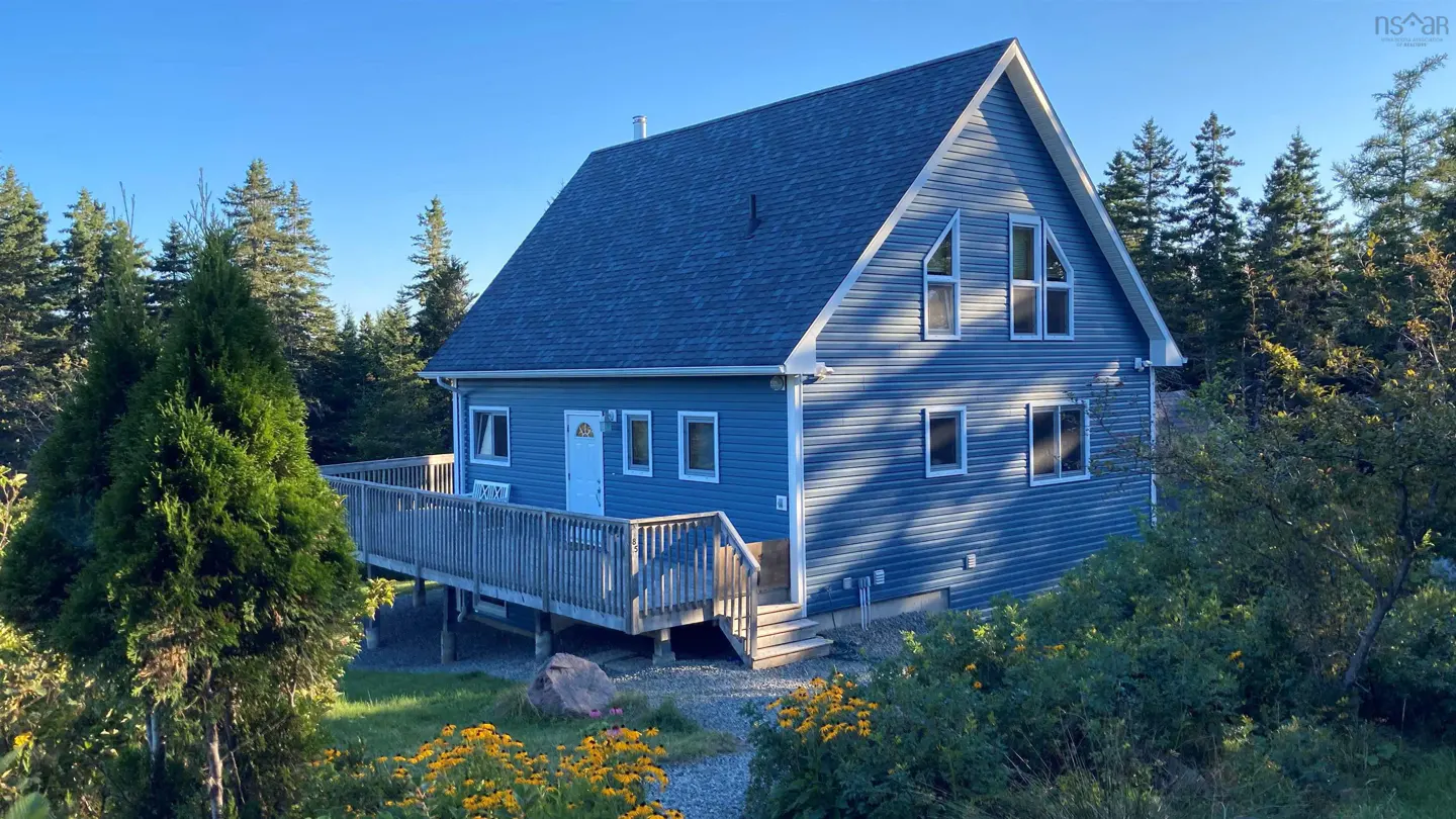 A blue, two-story house with a deck is surrounded by trees and yellow flowers.
