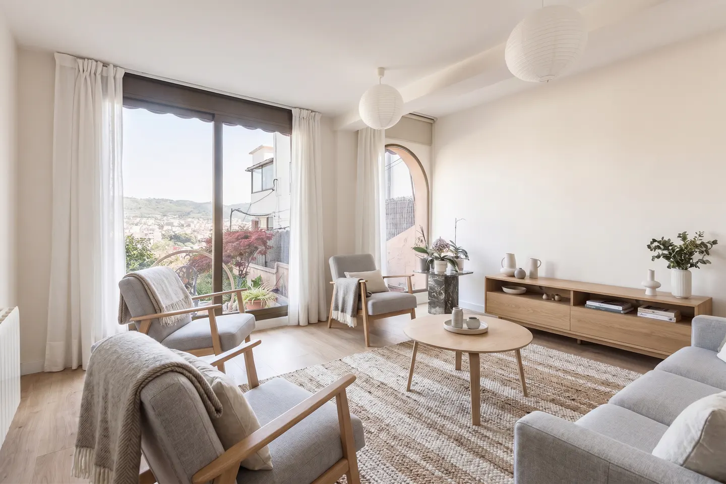 Bright living room with gray chairs, sofa, and wood furniture. Large window with a city view. White walls and paper lanterns.