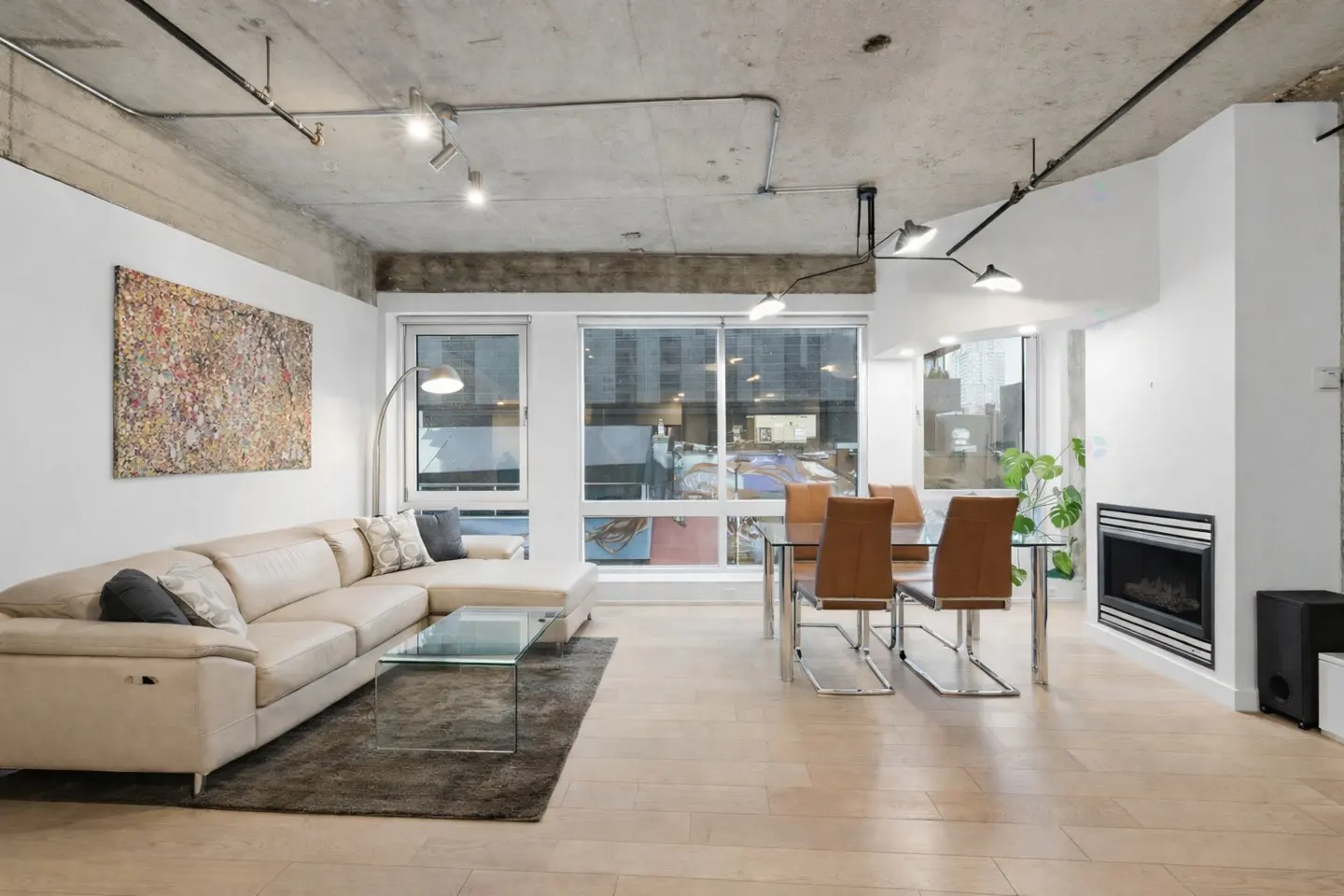 Bright living room with a beige sectional sofa, glass coffee table, and a dining table with leather chairs. A modern fireplace and large windows add to the space.