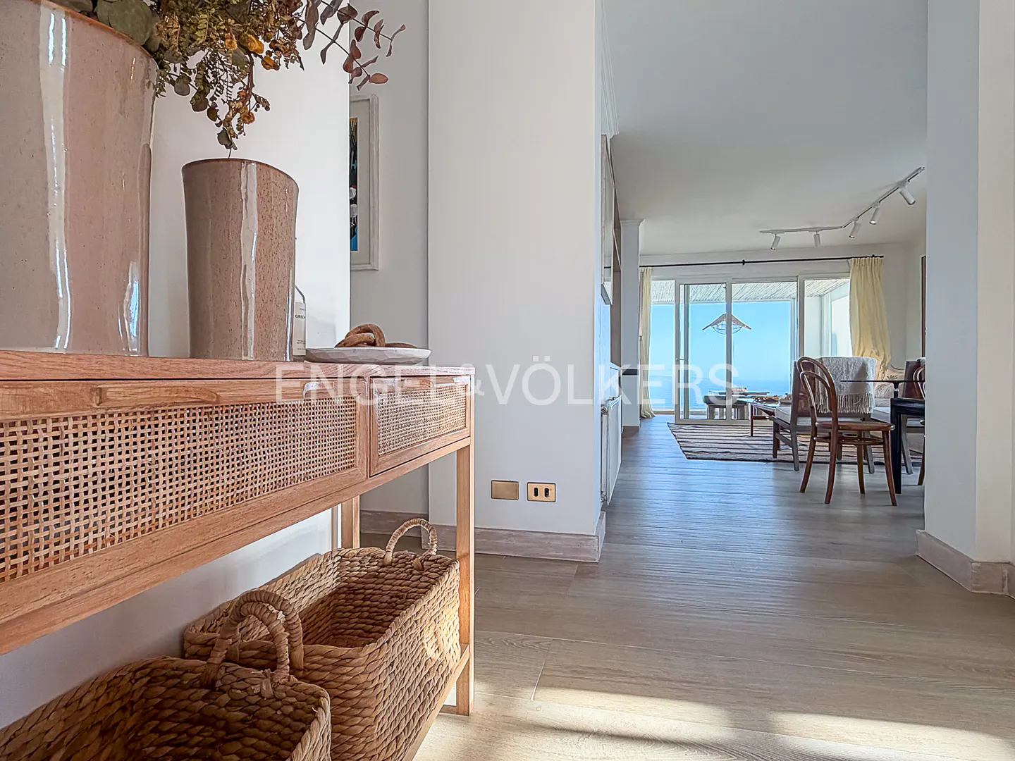 Entryway with a light wood console table, wicker baskets, and vases. Living room with ocean view in the background.