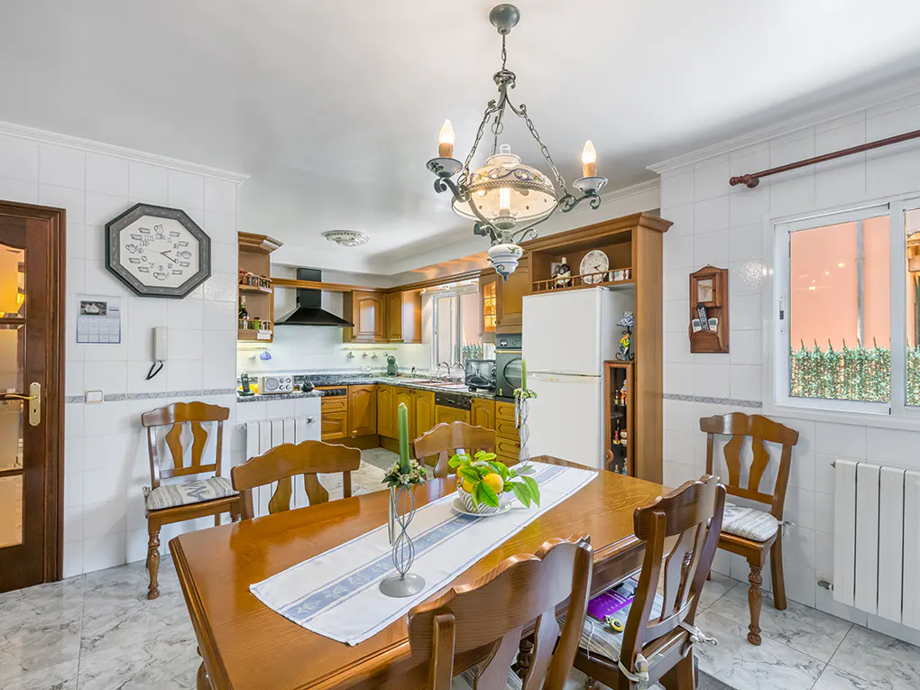 A bright kitchen and dining area with a wooden table, chairs, and a chandelier. The kitchen has wooden cabinets and white appliances.