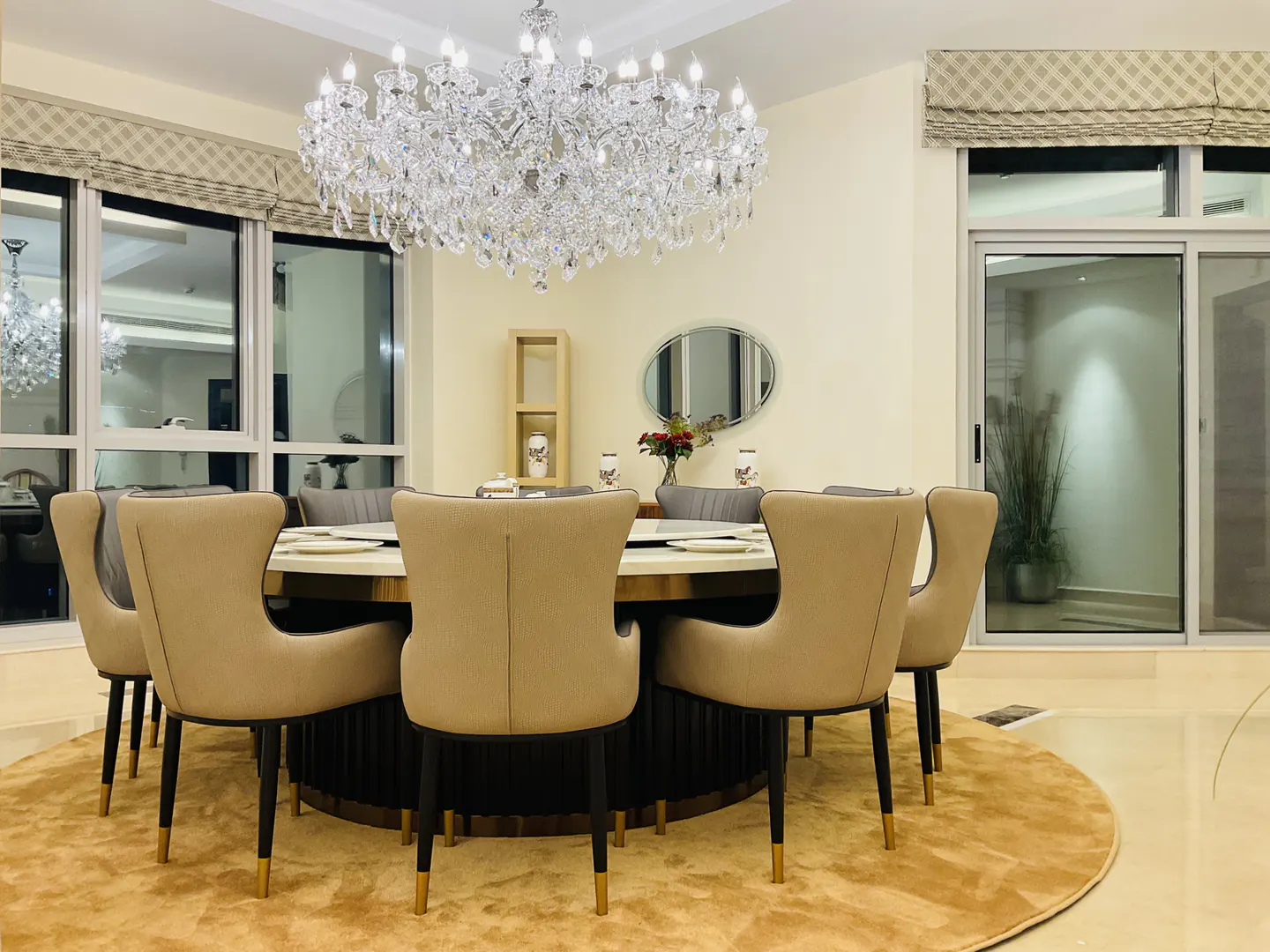 Dining room with a round table, beige chairs, and a large crystal chandelier. Windows with patterned blinds and a round rug complete the elegant space.