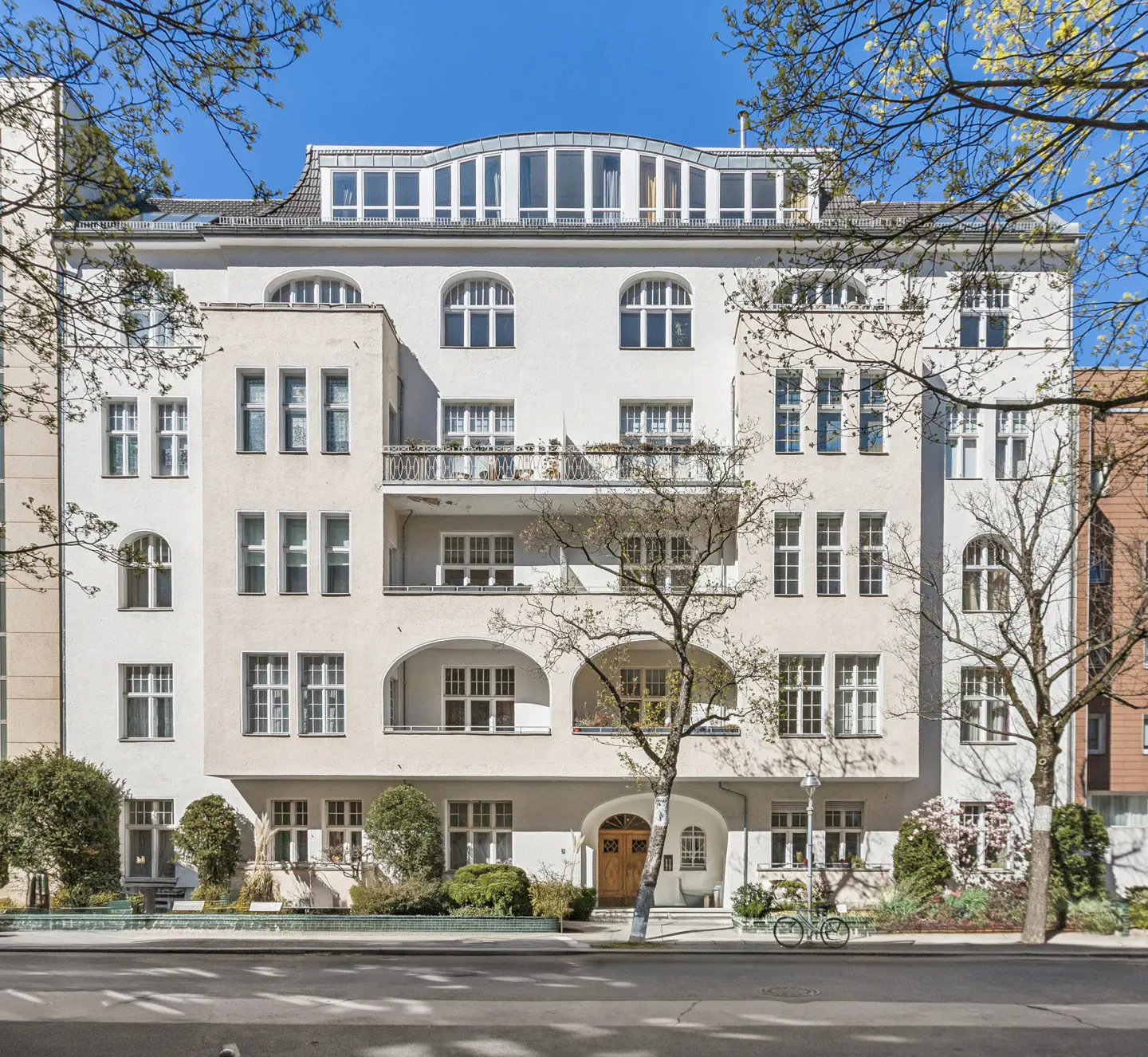 A light-colored, multi-story apartment building with arched windows and a balcony, framed by trees under a clear blue sky.