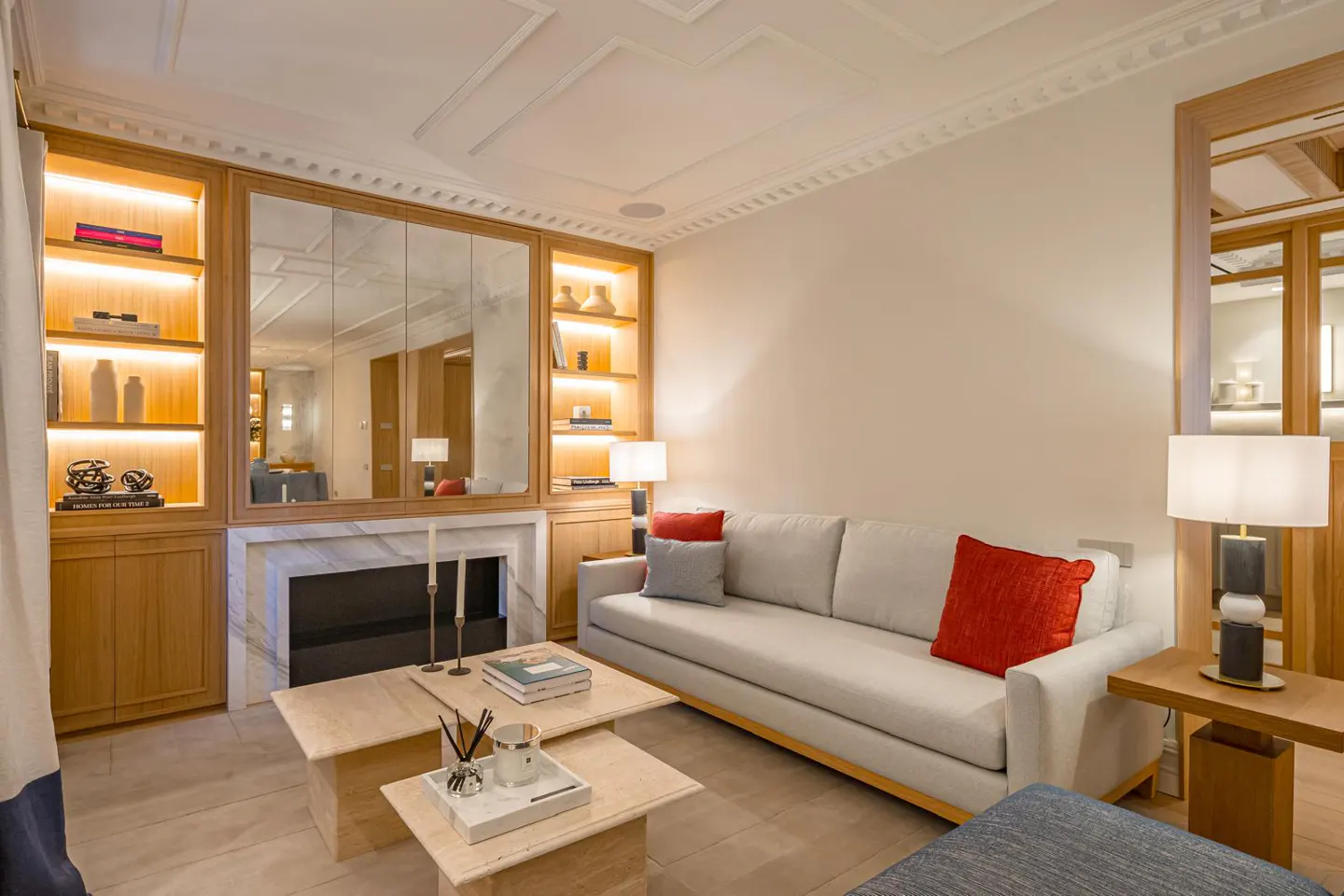 Living room with a light gray sofa, red pillows, and two square coffee tables. Built-in shelves with lighting flank a fireplace with a mirror above.