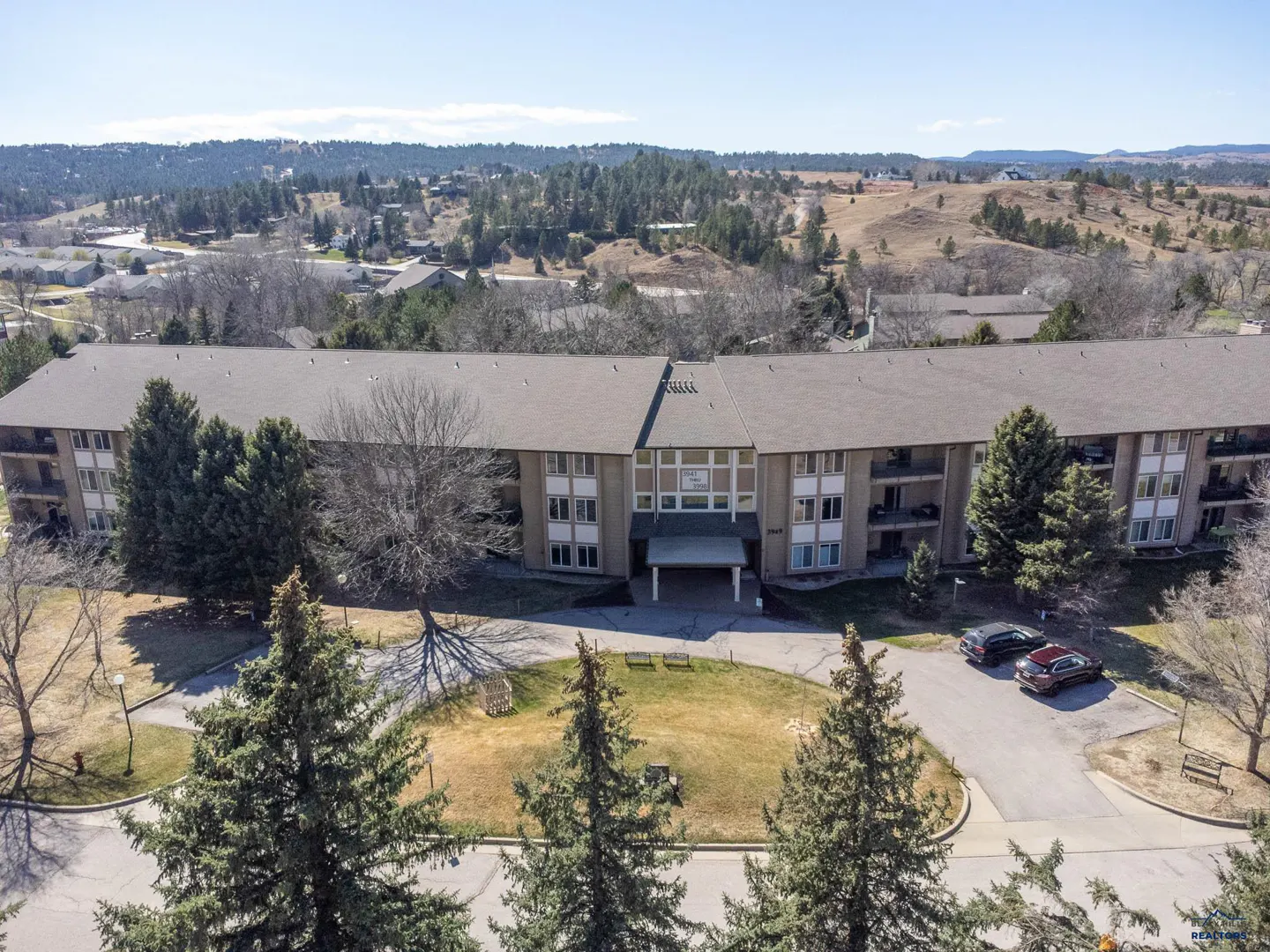 Aerial view of a three-story apartment building with a gray roof, surrounded by green trees and a grassy lawn. Two cars are parked in the driveway.