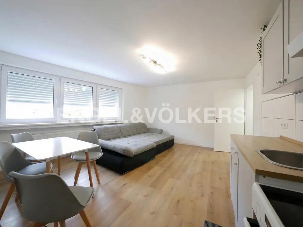 Bright open-plan living area with a grey sectional sofa, dining table with four chairs, and a modern kitchen corner.