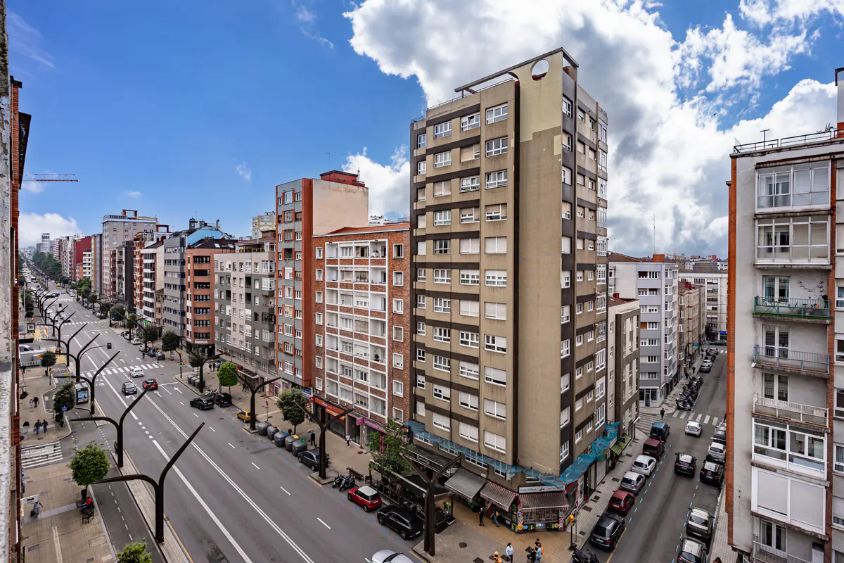 Cityscape view of a wide street lined with tall buildings, cars, and trees under a blue sky with clouds.