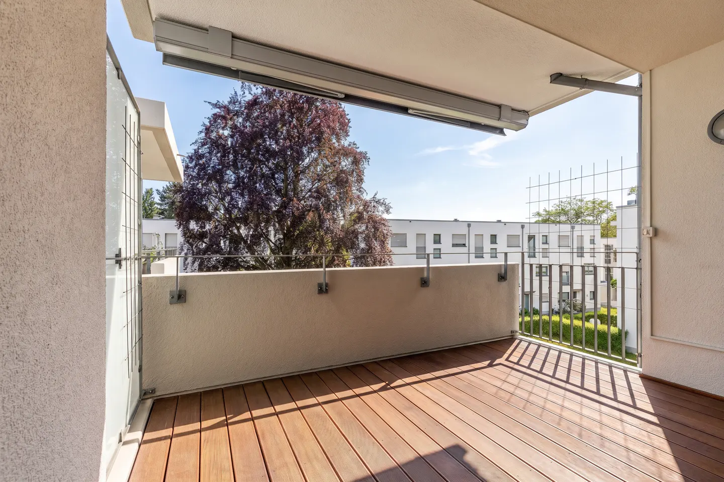 A covered balcony with wooden decking, a metal railing, and a view of a large tree and modern white apartment buildings.