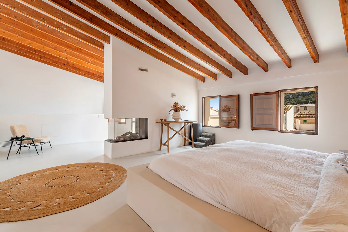 Modern bedroom with white walls, exposed wooden ceiling beams, a platform bed, a fireplace, and a round jute rug.