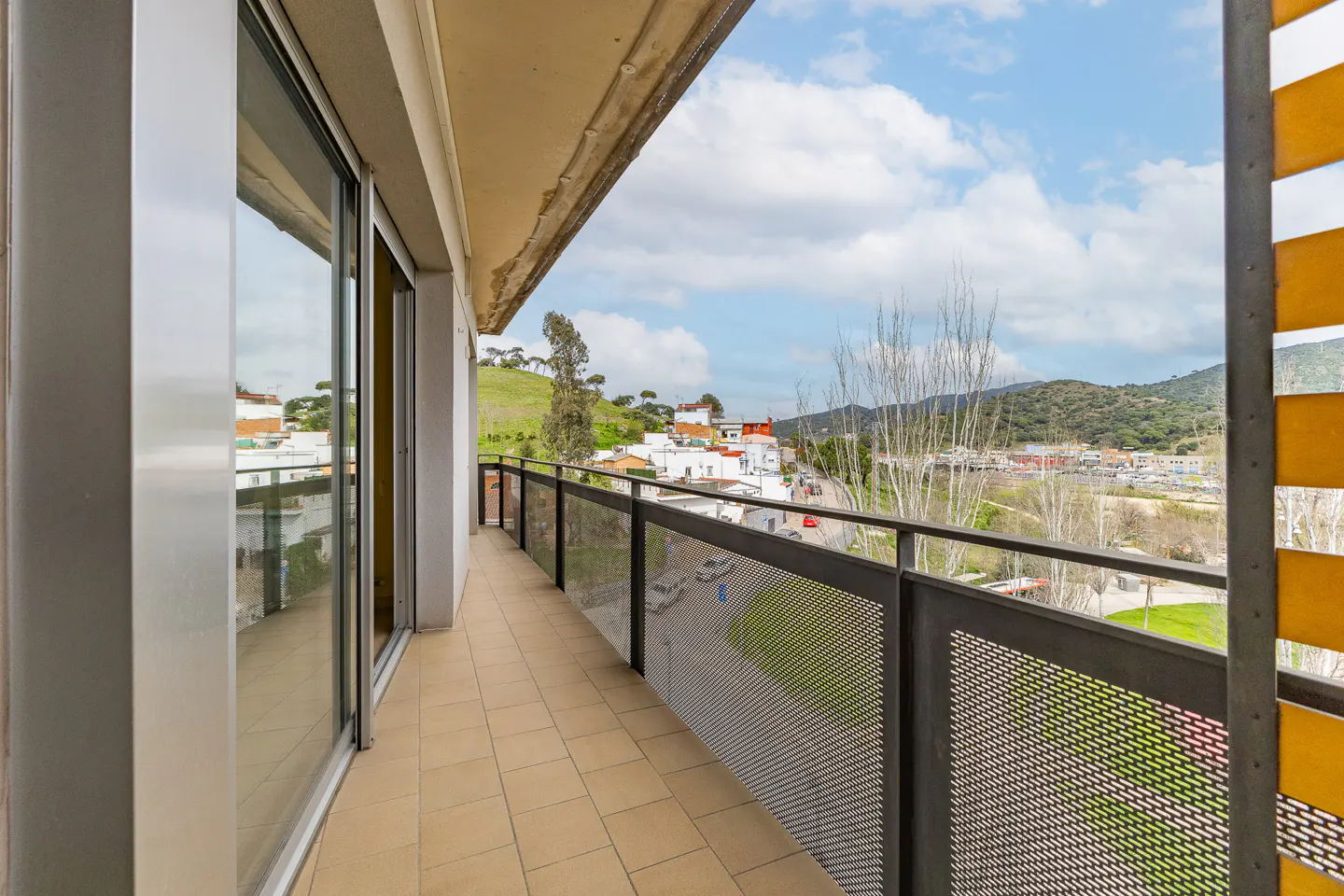 A balcony with beige tile flooring and a black metal railing overlooks a town with green hills in the background.