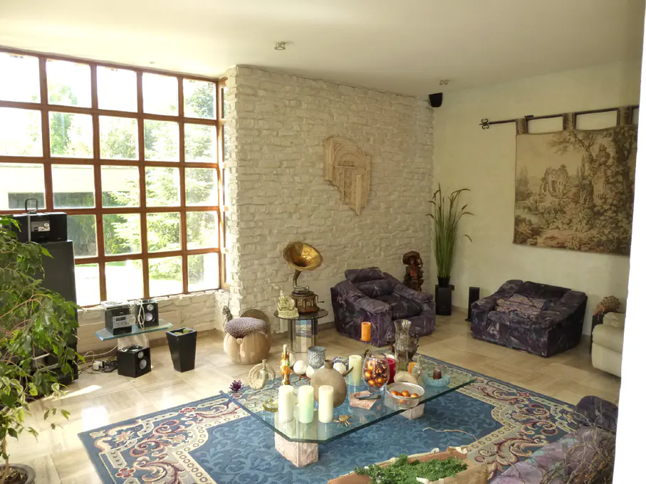 A spacious living room with a large window, stone wall, purple sofas, and a blue patterned rug.