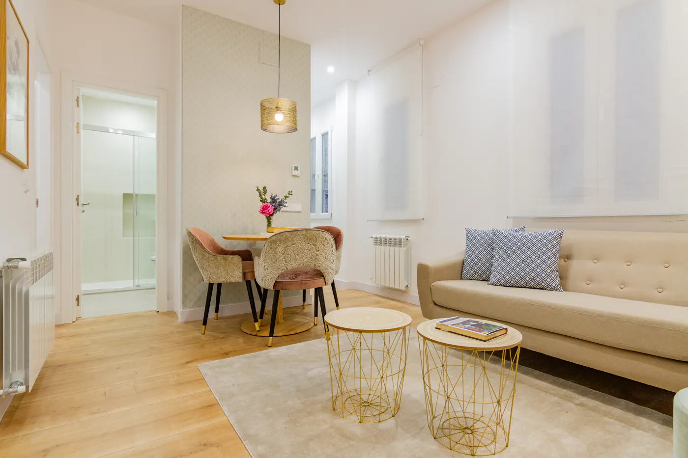 Bright living room with a beige sofa, patterned pillows, and two gold wire side tables. A round table with pink chairs sits under a gold pendant light.