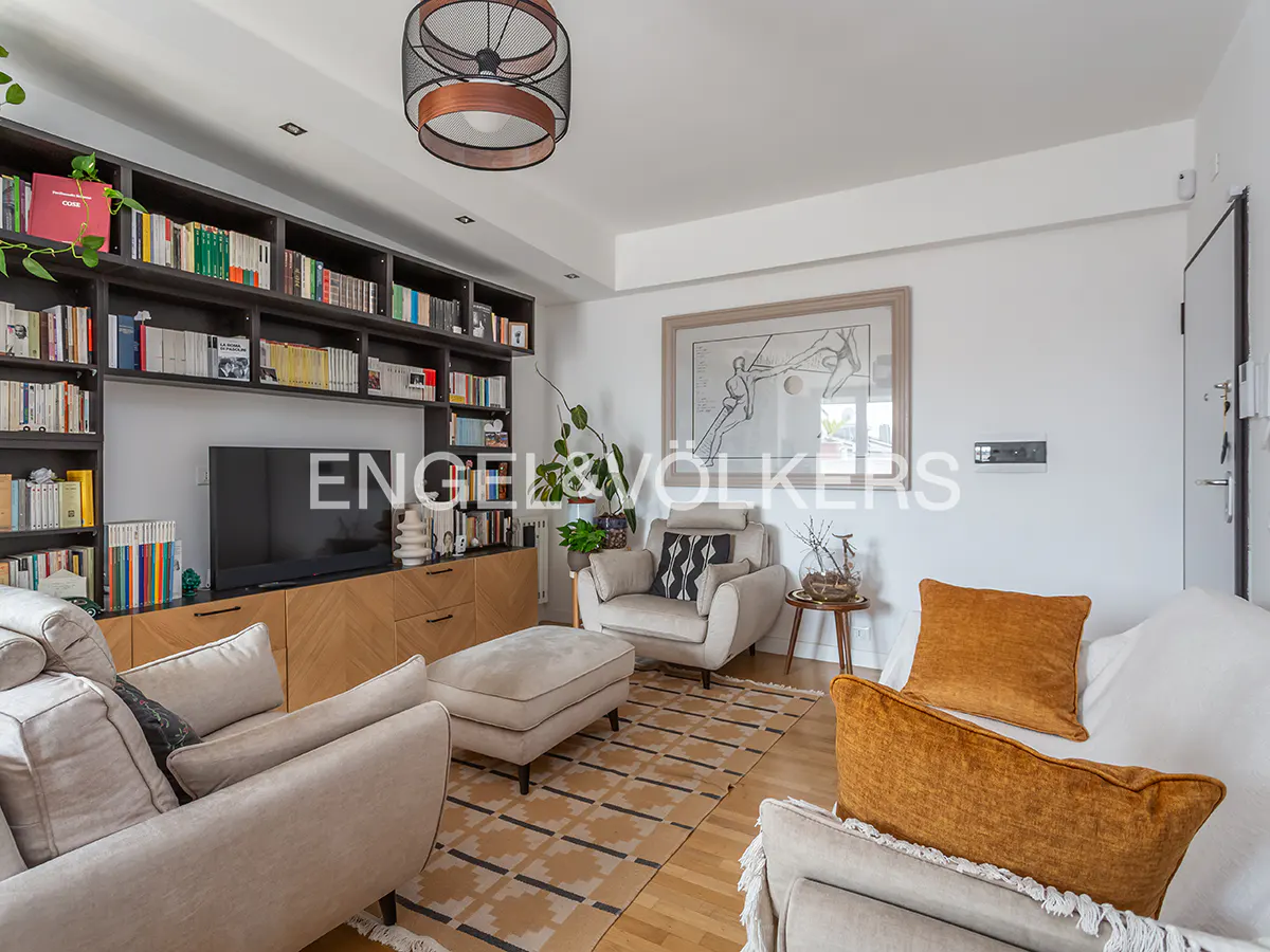 Modern living room with beige sofas, a patterned rug, a large bookshelf, a TV, and a framed line-art print on the wall.