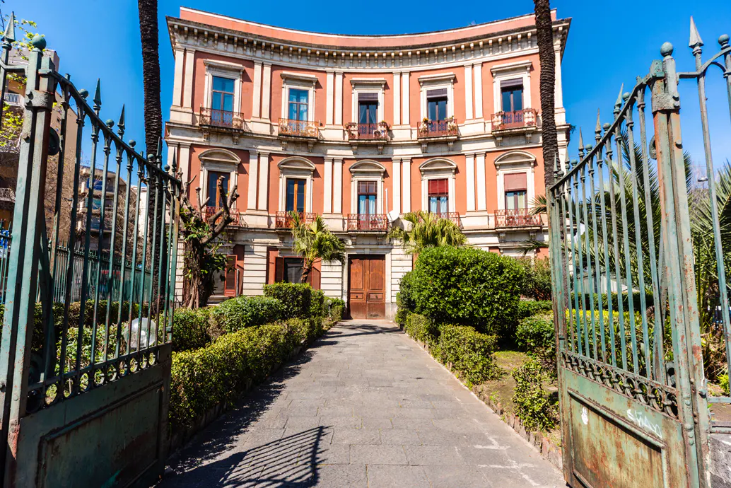 View of a three-story, orange building with a curved facade, seen through an open, green, wrought-iron gate. A stone path leads to the building's wooden door.