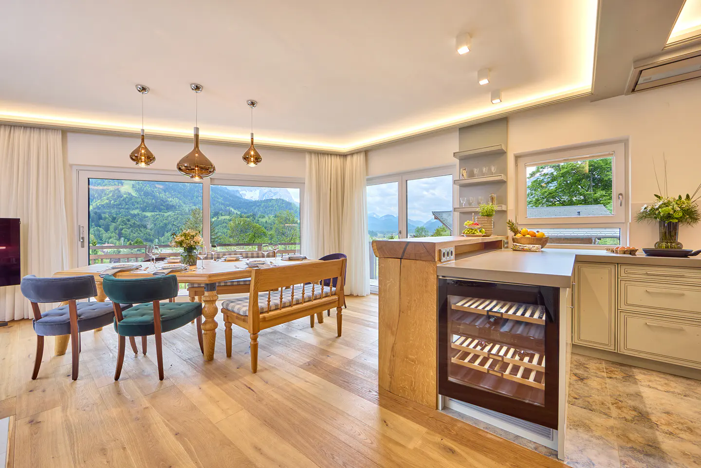 Bright, open kitchen and dining area with wood floors, table, chairs, and mountain views through large windows. Wine fridge visible.