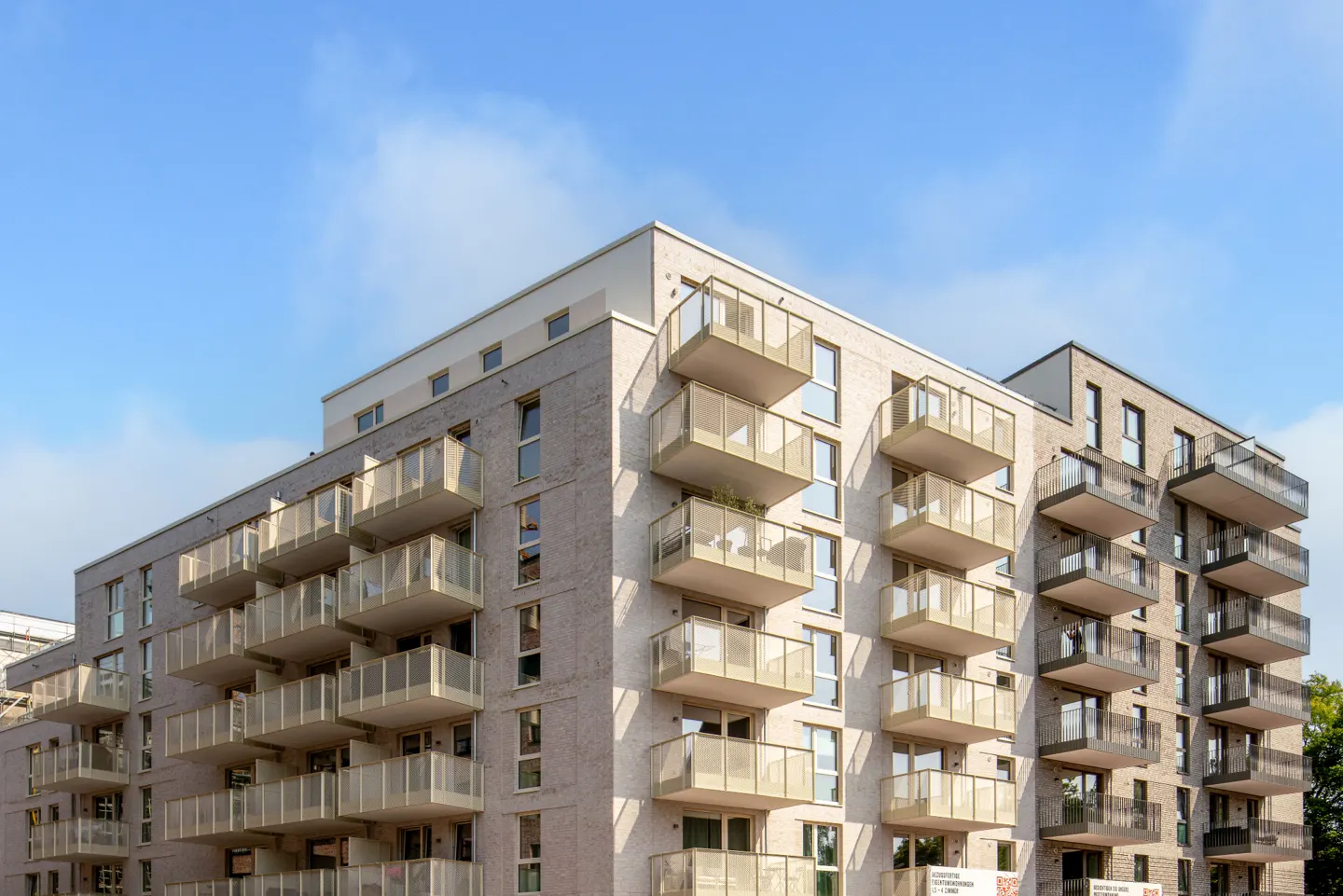Modern apartment building with light brick facade and balconies under a blue sky.