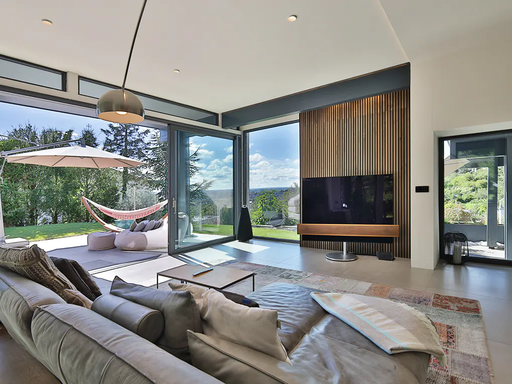 Bright living room with a gray sofa, large windows, and a view of a patio with a hammock and umbrella. A TV is mounted on a wood-paneled wall.