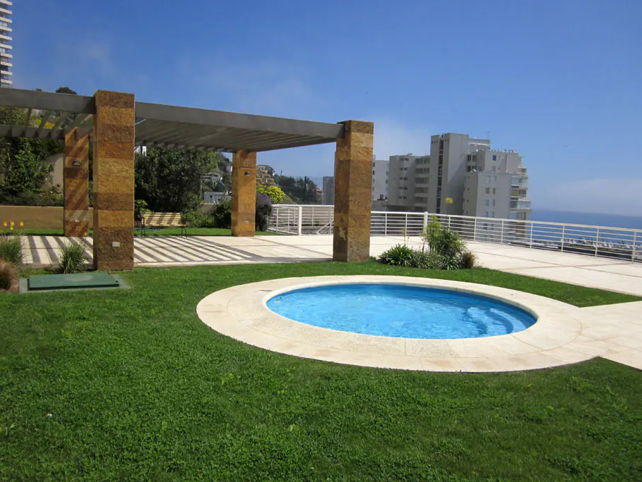 Outdoor patio with a small, round, blue pool surrounded by green grass, a stone pergola, and a view of the ocean and buildings.