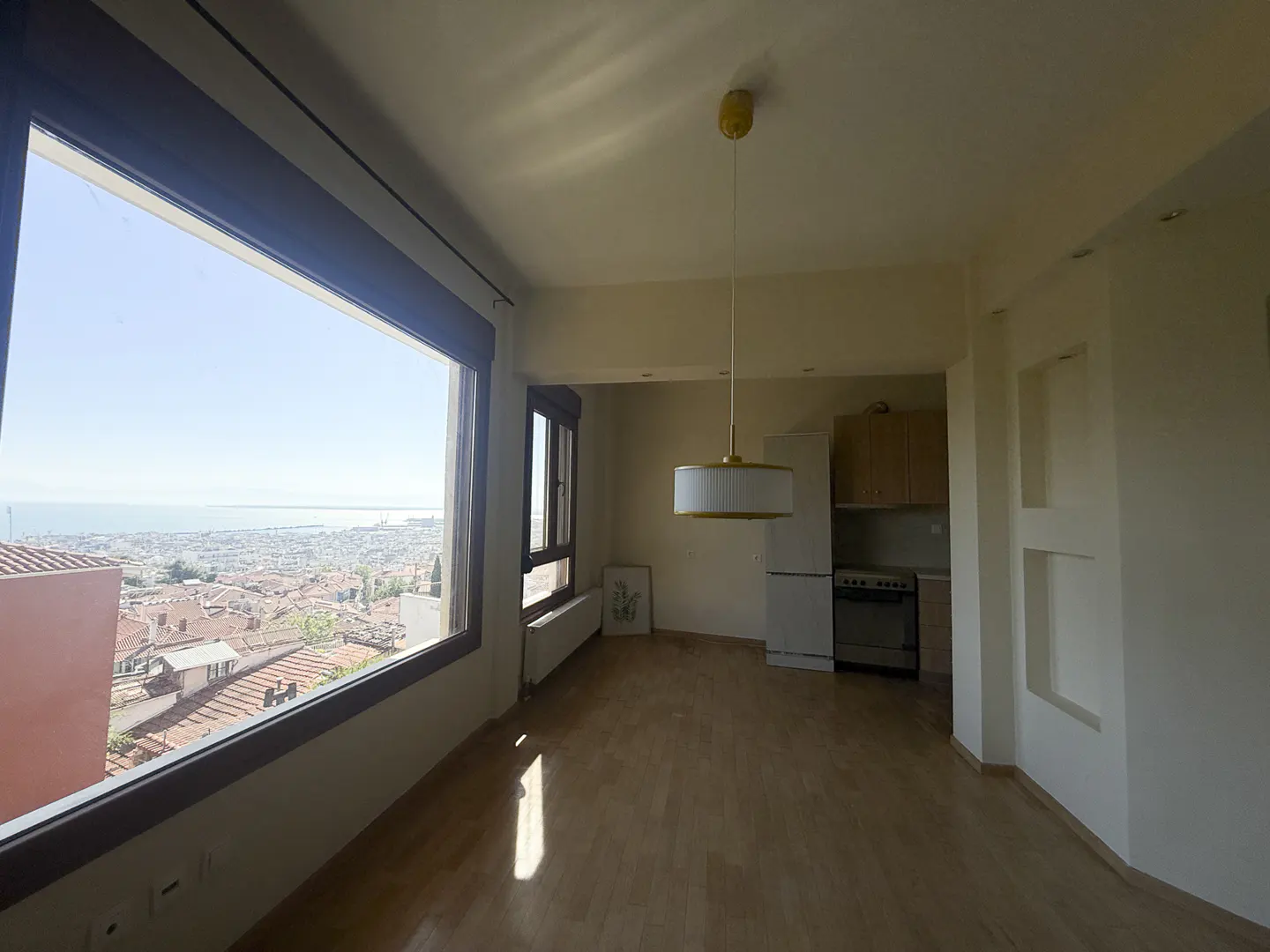 Bright room with hardwood floors, a large window overlooking a city and sea, and a small kitchen area with a pendant light.