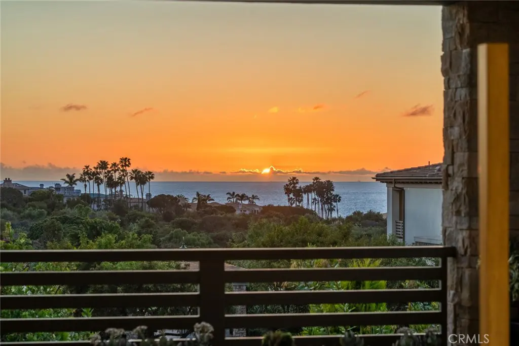 View from a balcony with a brown railing overlooking the ocean at sunset. Palm trees and greenery are visible in the distance.