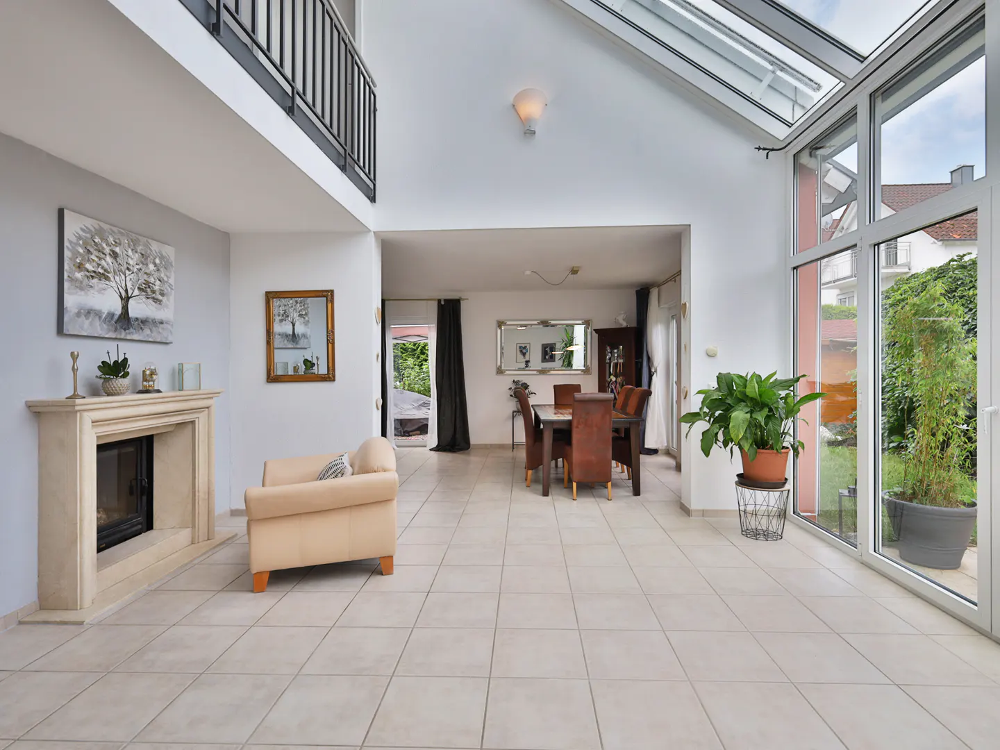 Bright, open-plan living room with a fireplace, beige sofa, dining table, and floor-to-ceiling windows overlooking a garden.