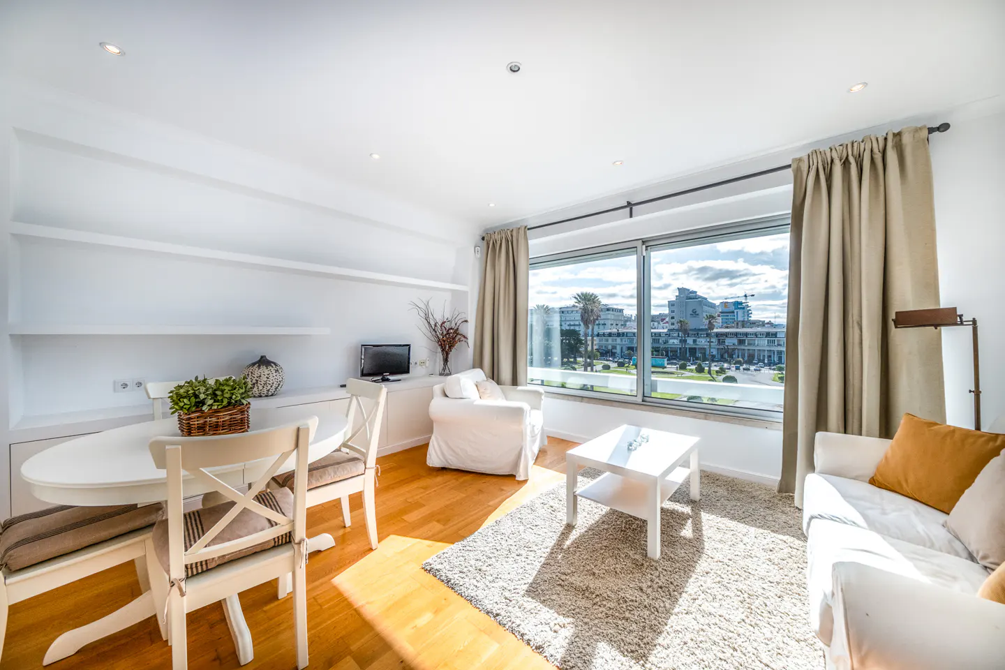 Bright, modern living room with white furniture, wood floors, and a large window overlooking a city. A dining table and chairs are visible.