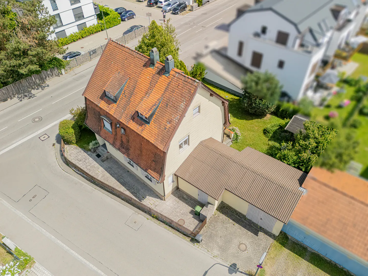 Aerial view of a cream-colored house with a red tile roof and attached garage, surrounded by green grass and trees.
