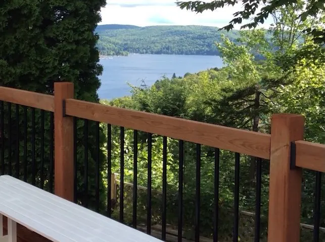 View from a deck with a wooden railing and black metal spindles overlooking a lake surrounded by green trees and hills.