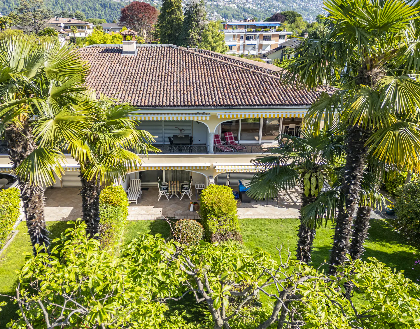 Two-story house with a red tile roof, yellow trim, and palm trees in the yard. Balconies with furniture overlook the green lawn.