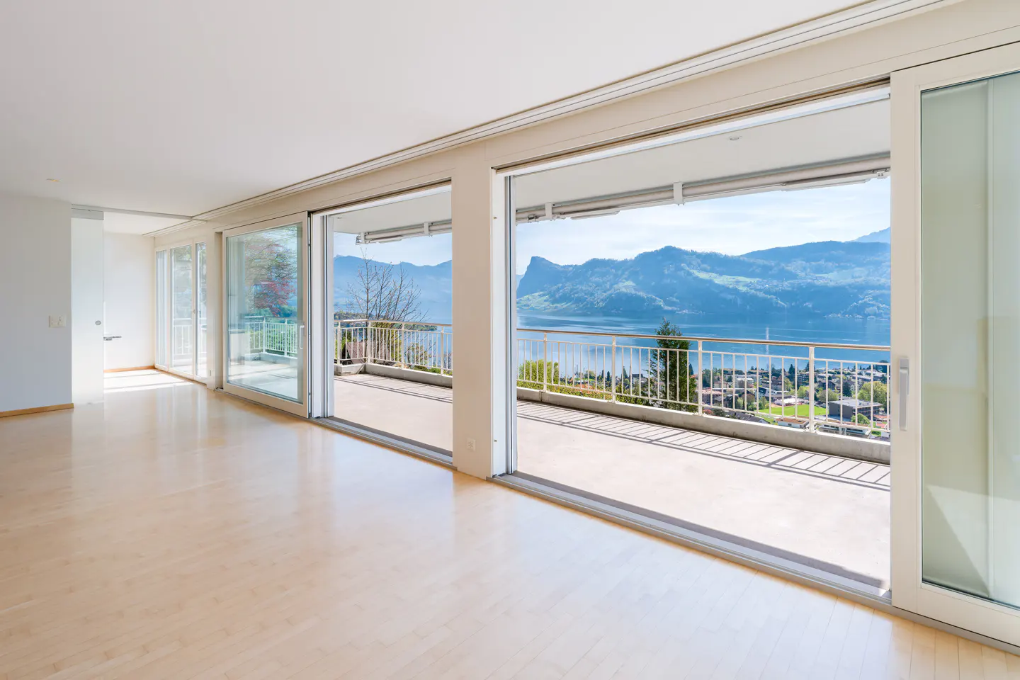 Bright, empty room with light wood floors and sliding glass doors to a balcony overlooking a lake and mountains.