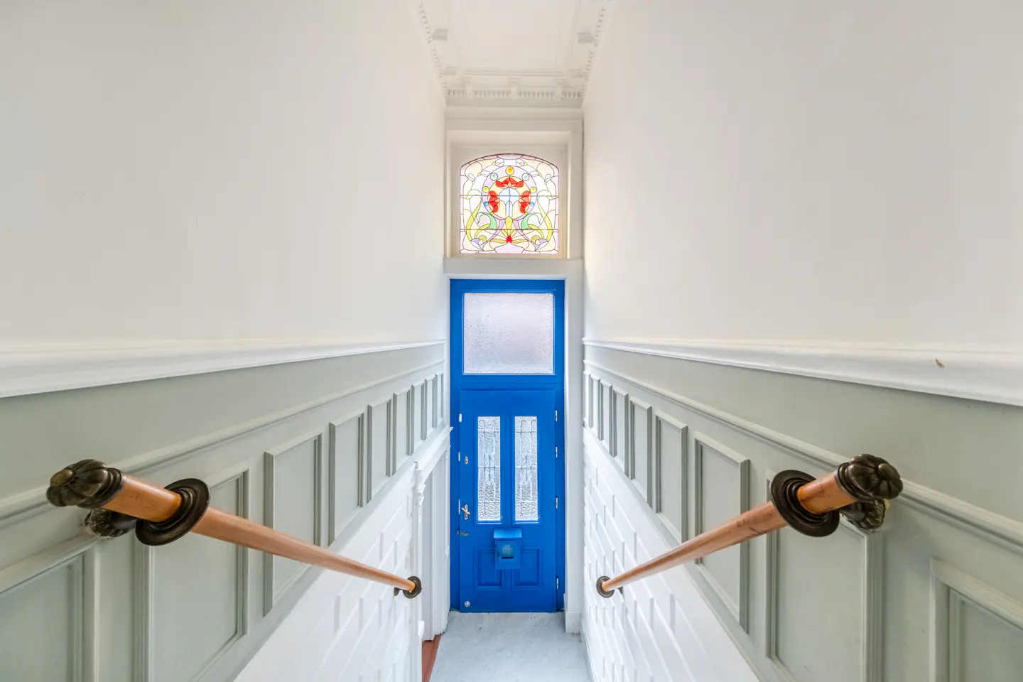 Hallway view with white walls, gray wainscoting, and wooden handrails. A bright blue door with a stained glass window above is at the end of the hall.