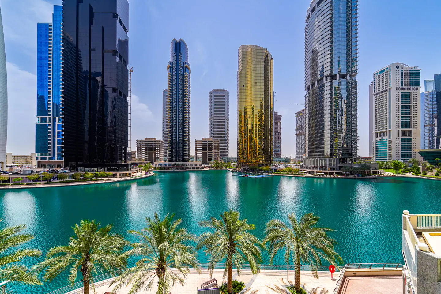 View of Dubai skyscrapers surrounding a turquoise lake, with palm trees in the foreground.