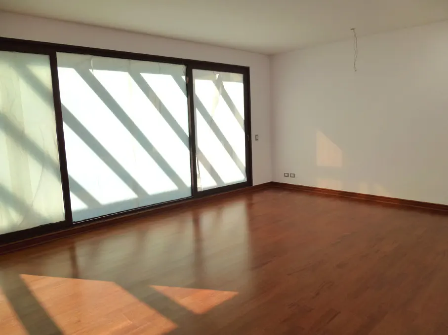 Empty room with hardwood floors, white walls, and large windows with sheer curtains letting in sunlight.