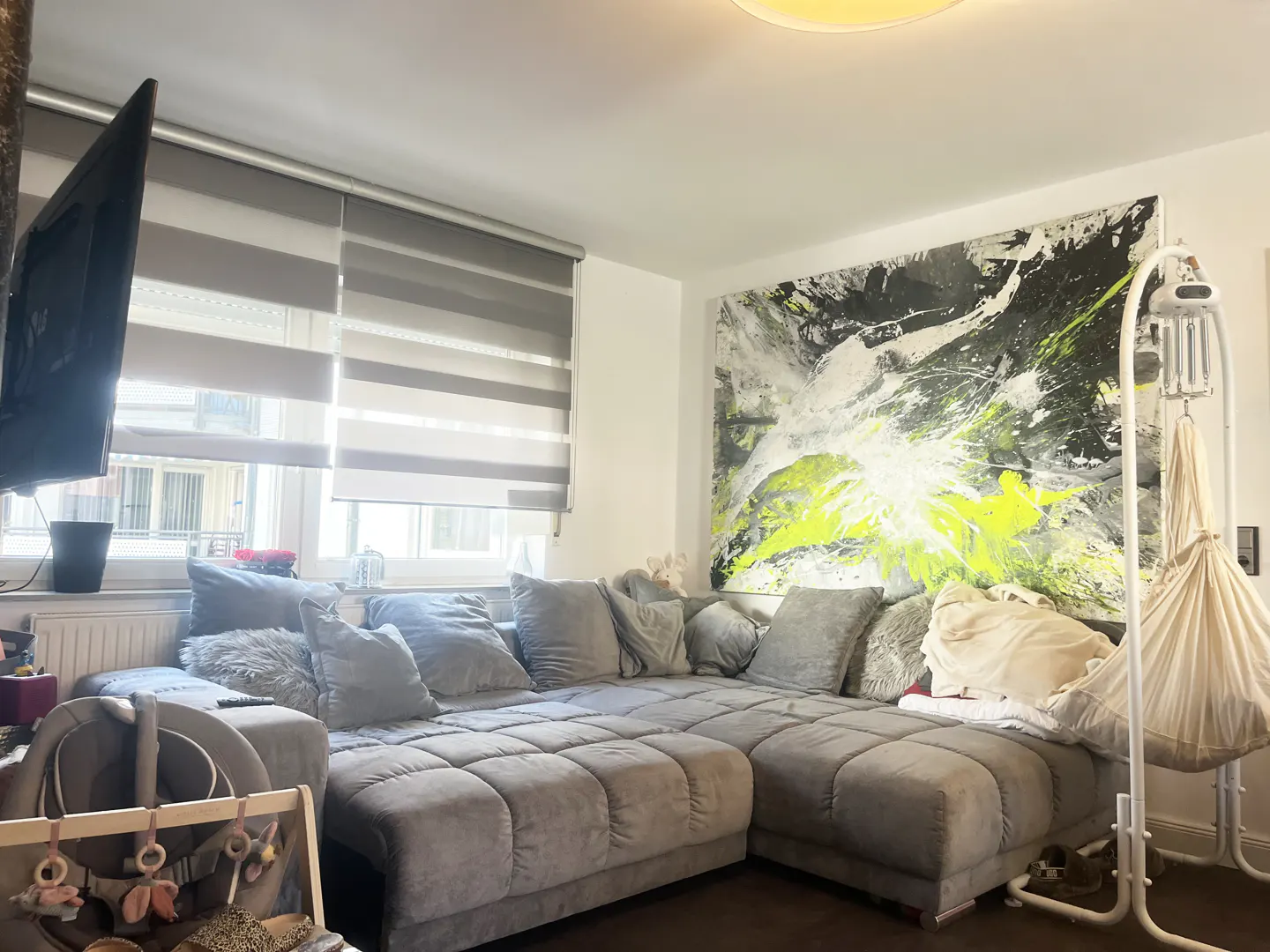 Living room with a large gray sectional sofa, abstract art with neon green accents, and a baby hammock. Window with blinds.