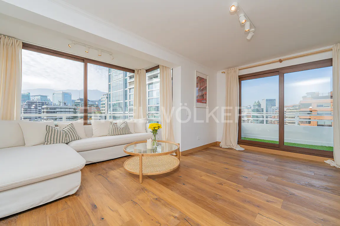Bright living room with wood floors, white sofa, and city view through large windows. A round glass coffee table sits in the center.