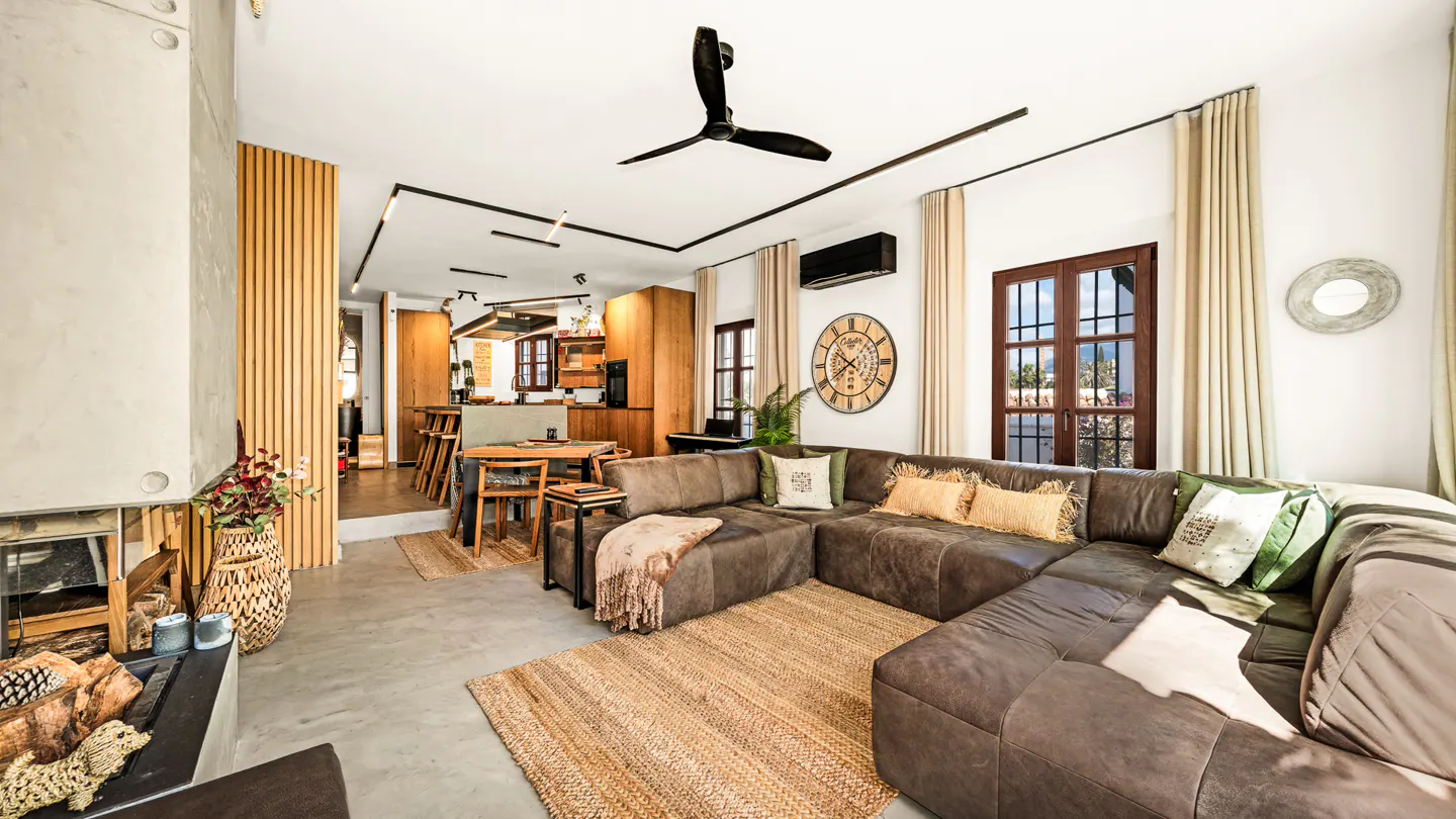 Open-concept living space with a brown sectional sofa, jute rug, and wood accents. A kitchen is visible in the background.
