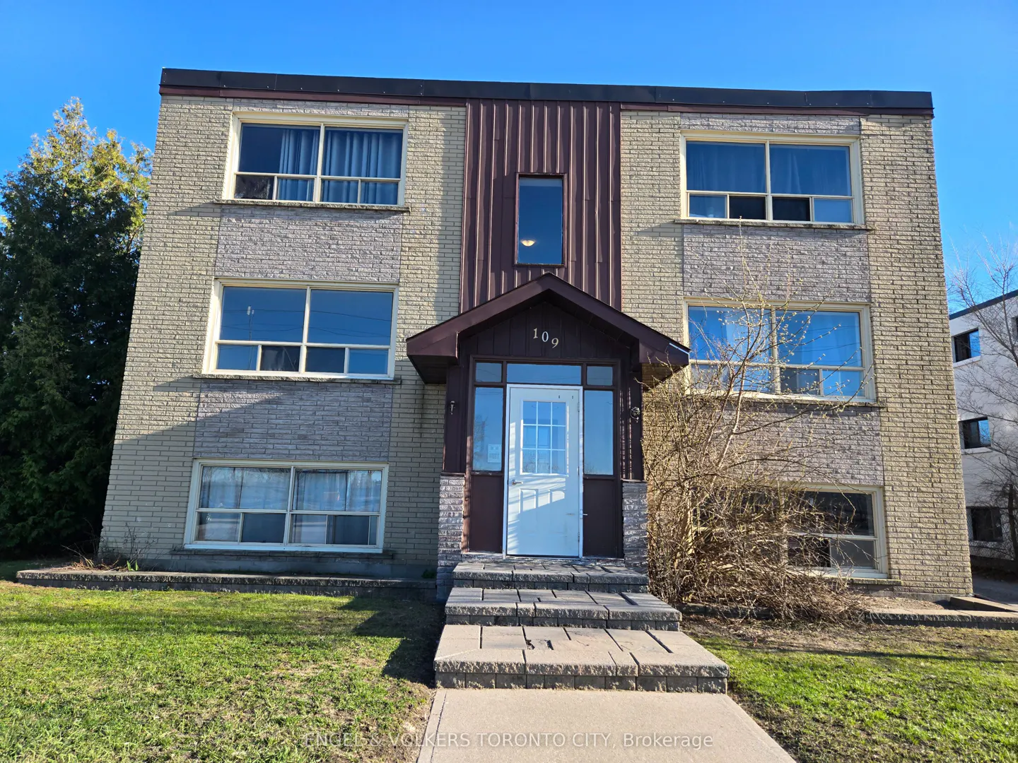 Three-story brick apartment building with a central brown entryway, white door, and stone steps under a clear blue sky.
