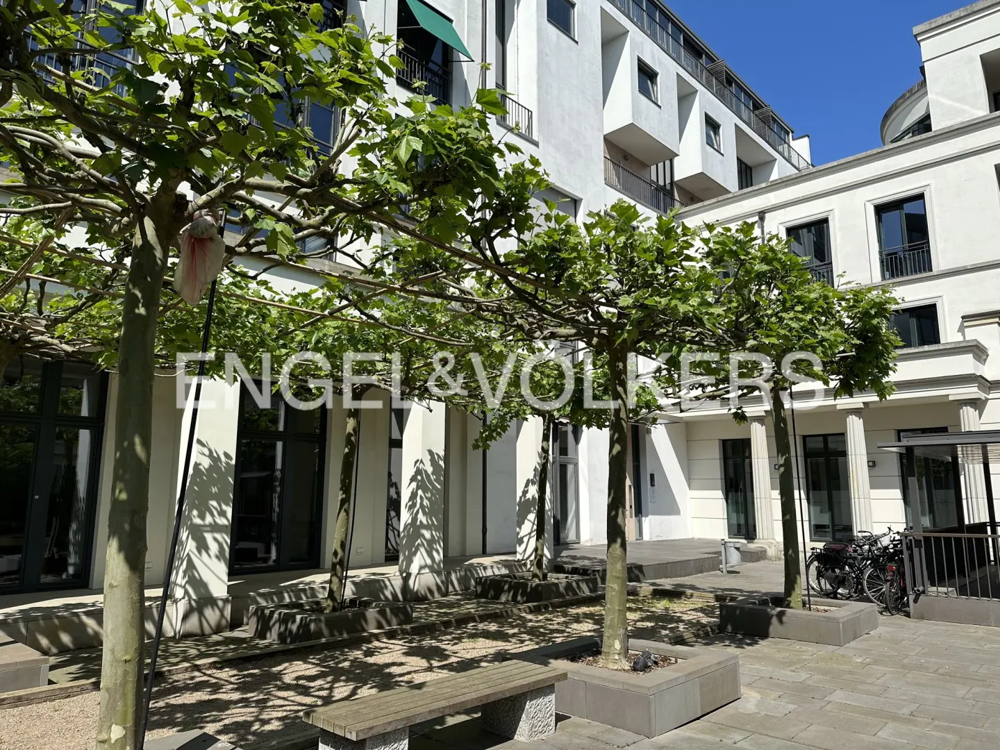 Exterior view of a white building with trees, benches, and the Engel & Volkers logo. Bicycles are parked near the building.