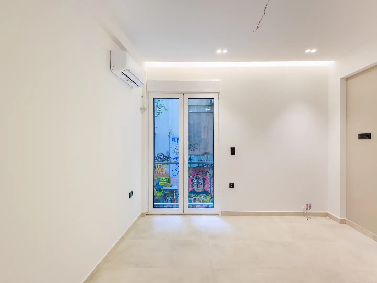 Bright, empty room with white walls, beige tile floor, and white double doors with graffiti art leading outside.