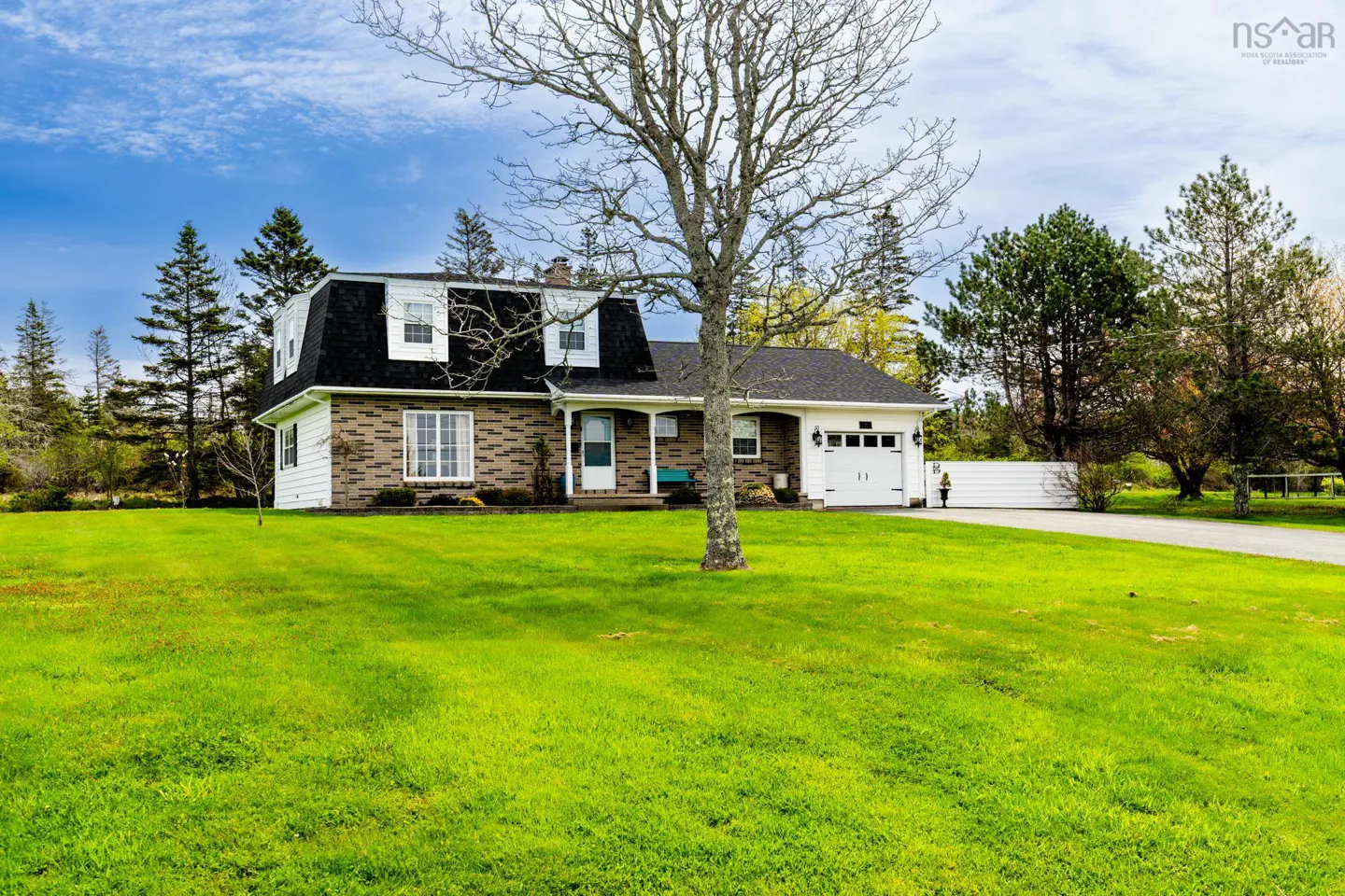 A brick house with a black roof and white garage door sits on a large green lawn under a blue sky.
