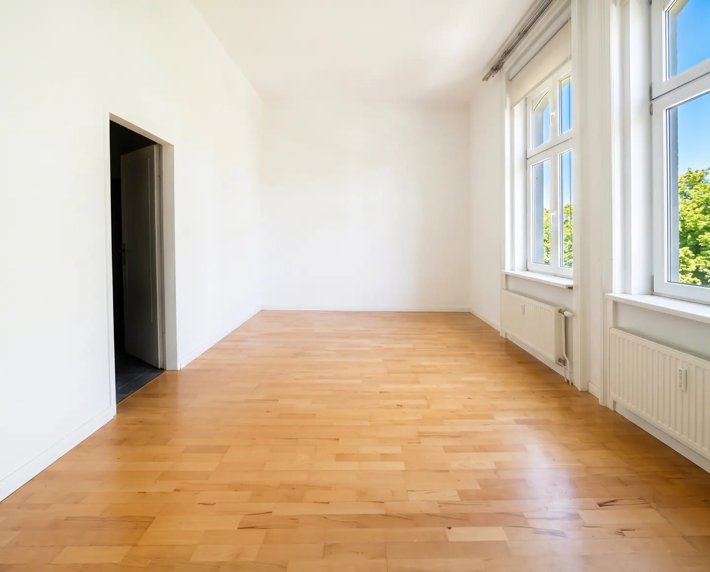Bright, empty room with light wood floors, white walls, and two large windows showing green trees. An open doorway is on the left.