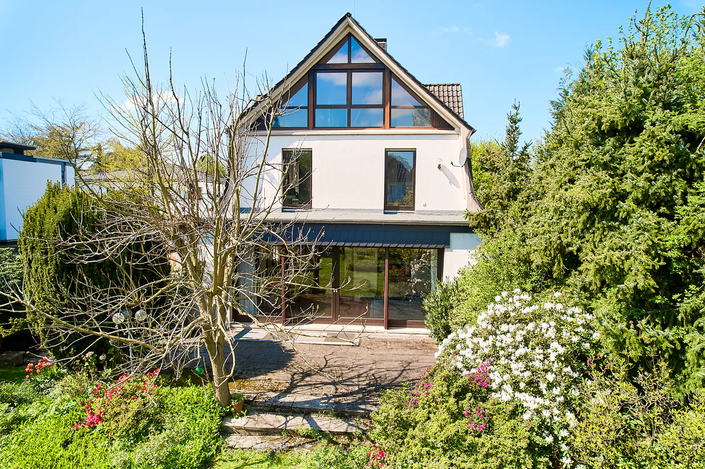 Two-story white house with a peaked roof and large windows, surrounded by lush greenery and flowers under a blue sky.