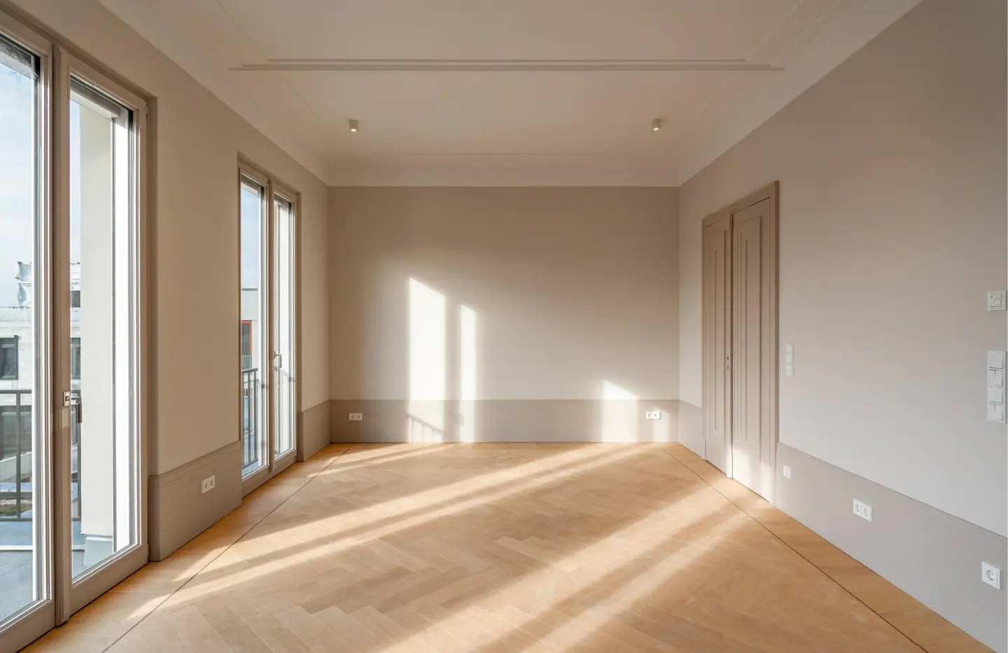 Empty room with light beige walls, herringbone wood floors, and tall glass doors letting in bright sunlight.