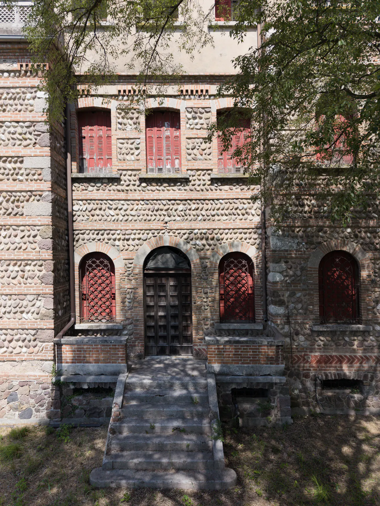 Stone building facade with arched dark wooden doors, red shuttered windows, and stone steps leading to the entrance.