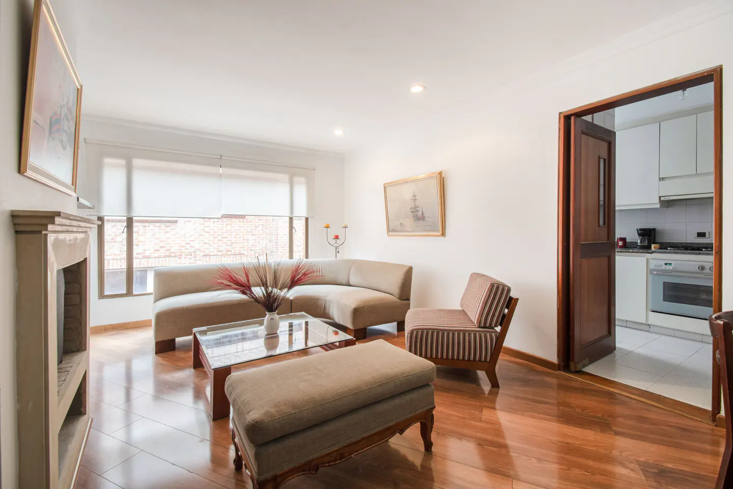 Living room with hardwood floors, beige sofa, striped chair, glass coffee table, and a view into a white kitchen.