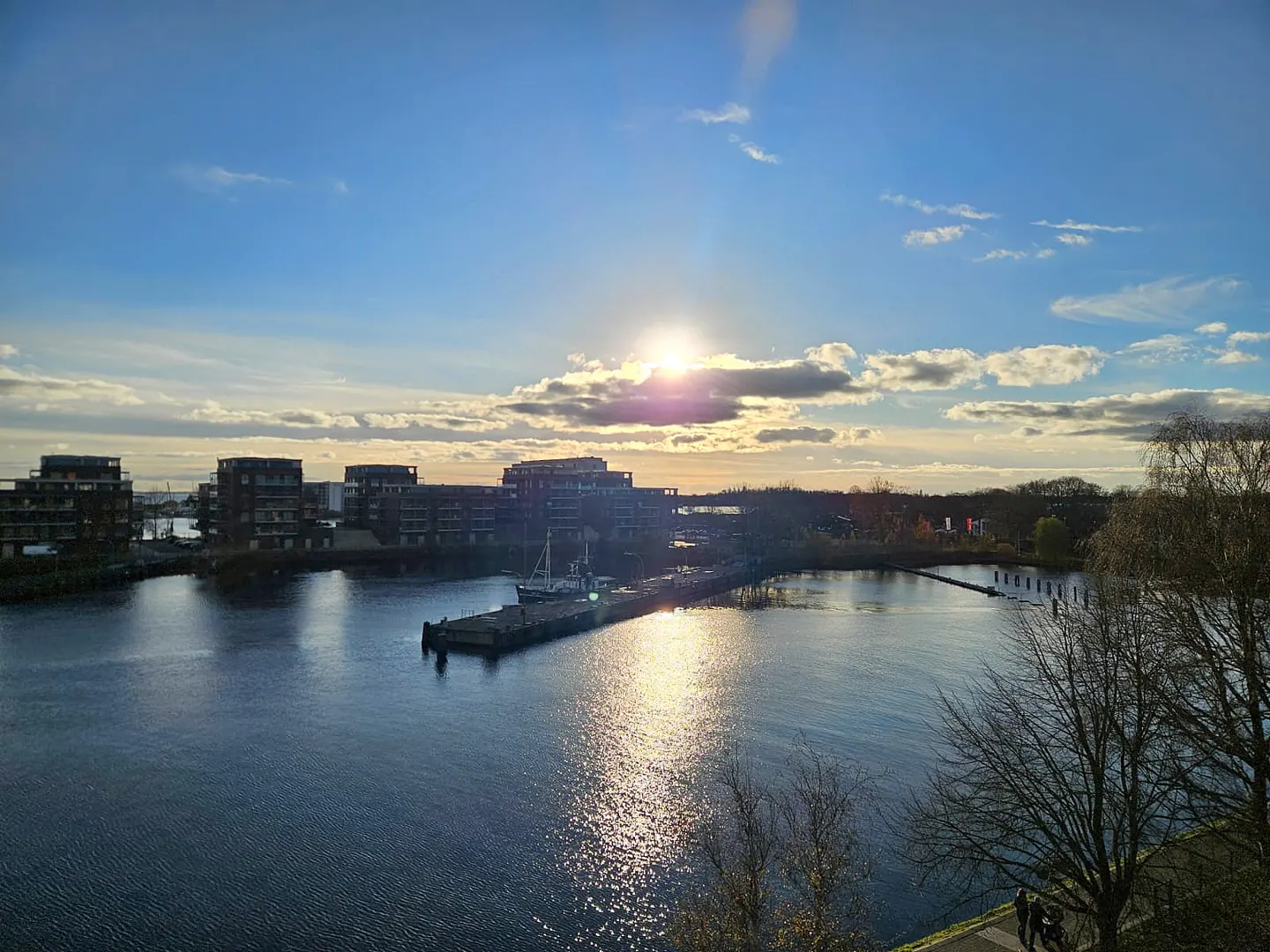Waterfront view of modern buildings, a pier with a boat, and a blue sky with the sun shining through clouds.