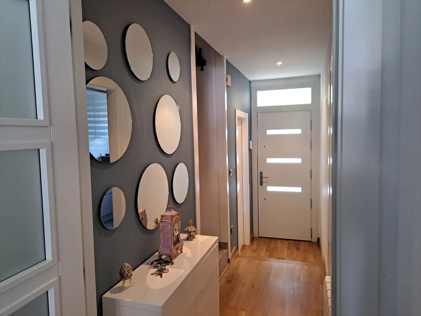 Hallway with gray walls, white trim, and wood floors. A white console table sits beneath a wall decorated with round mirrors. A white front door is at the end of the hall.
